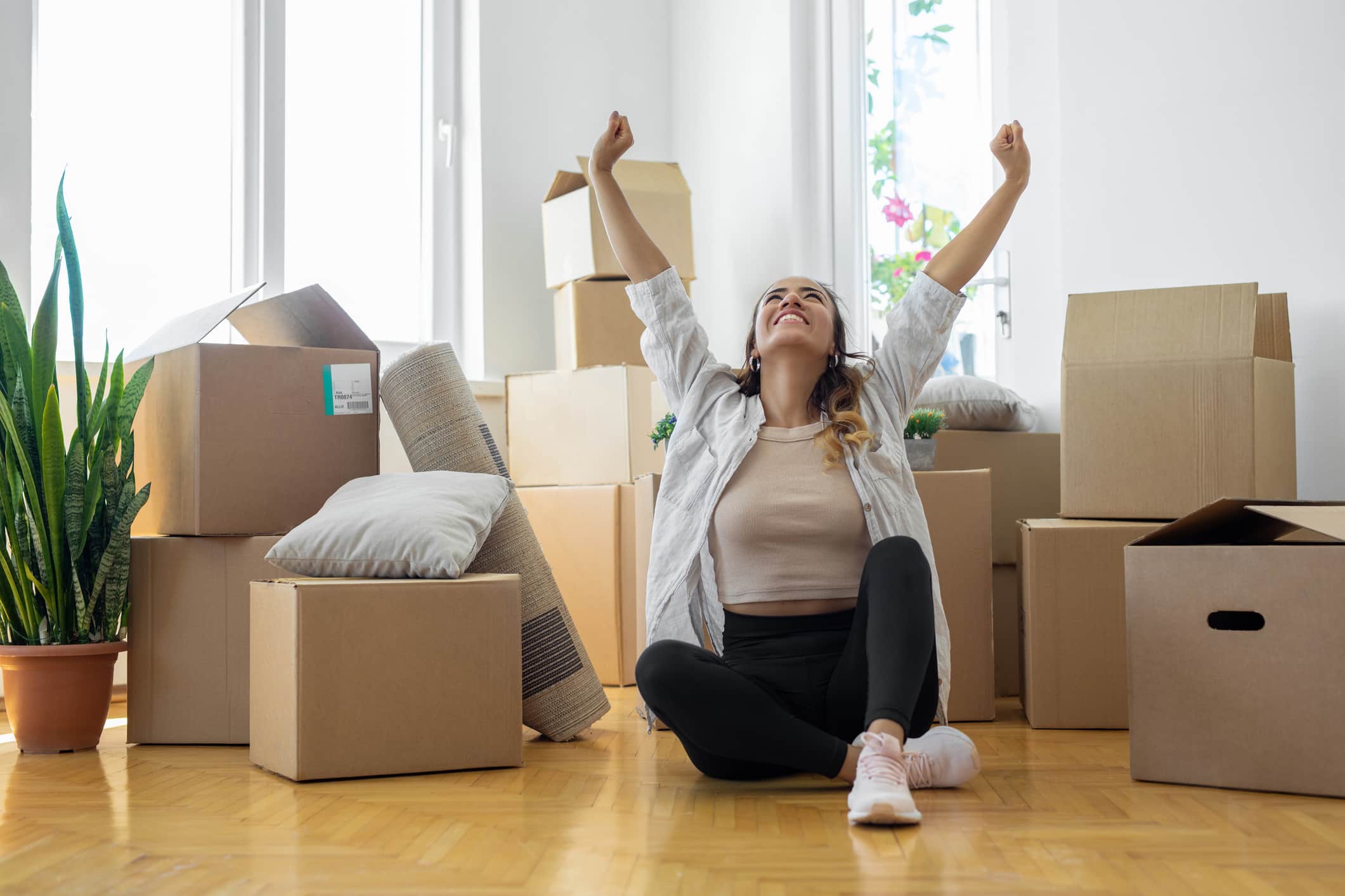 Young woman sitting on floor in new apartment with boxes and raising arms in joy